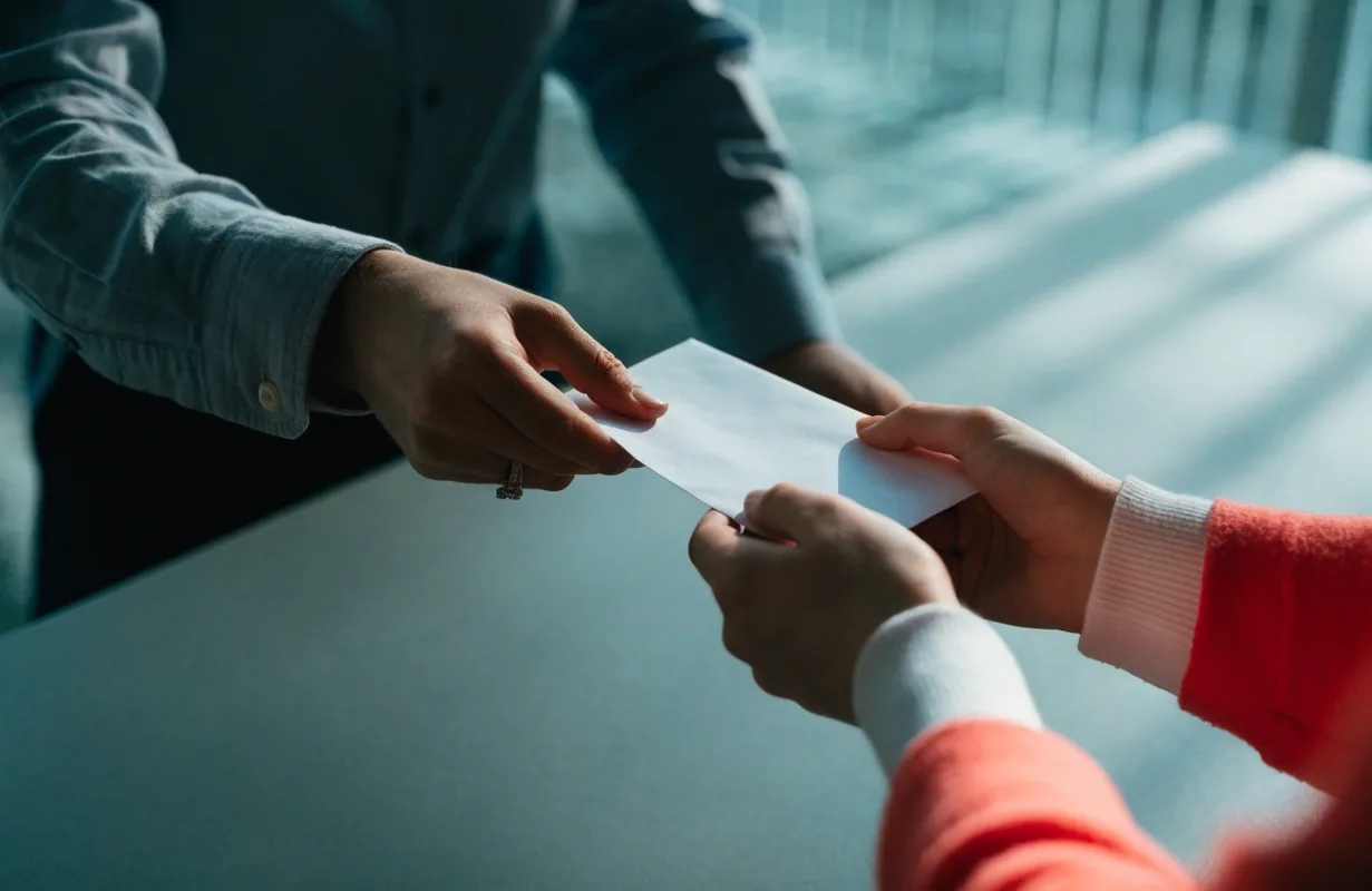 Two hands exchanging an unmarked envelope across a matte desk in cool daylight